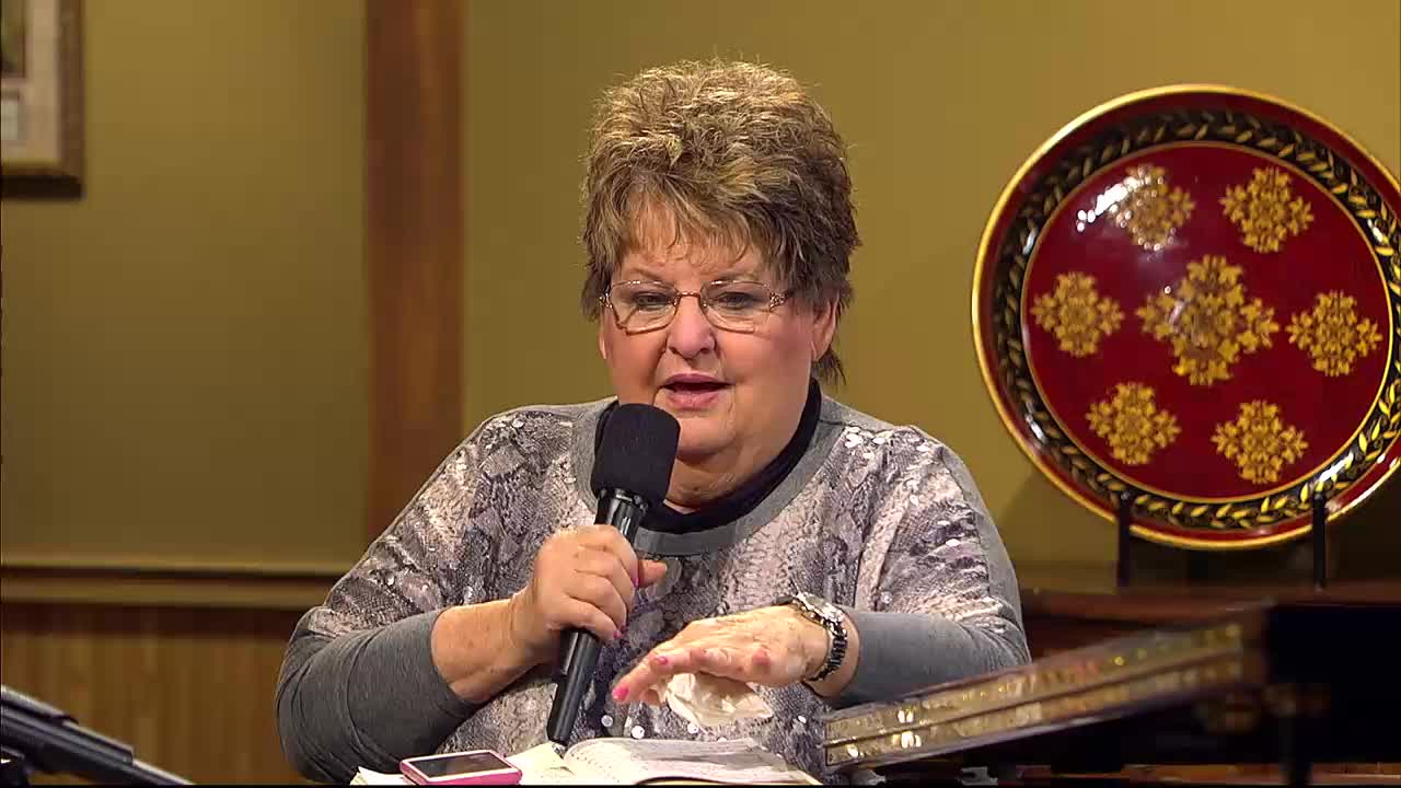 A woman speaks into a microphone, her hand gesturing over an open Bible. A decorative plate hangs on the wall behind her.