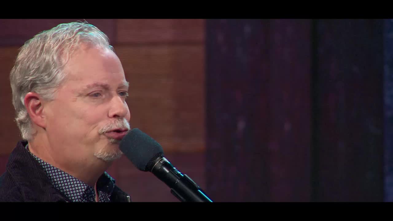 A man with graying hair and a mustache speaks into a microphone. He's wearing a dark jacket over a patterned shirt, likely on a program for the 3ABN Praise Him Music Network.