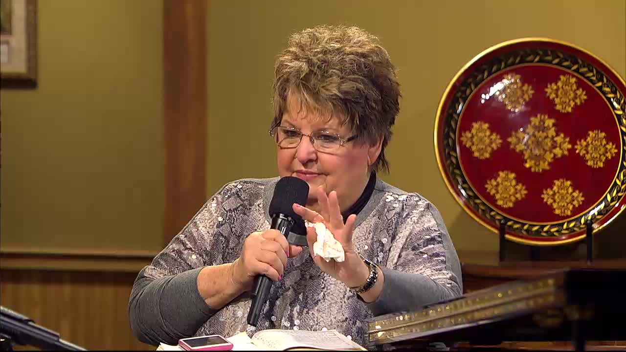 A woman with short, dark hair and glasses holds a microphone in her left hand and a crumpled tissue in her right. She speaks into the microphone, her expression earnest, as a large, ornate red plate sits on a stand behind her. This scene unfolds during a broadcast for the 3ABN Praise Him Music Network.