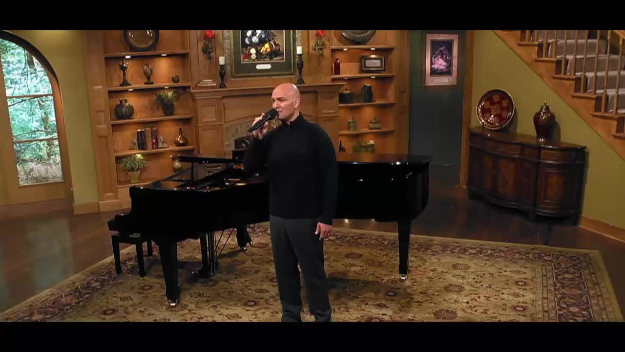 A man in a black sweater and dark pants stands before two grand pianos, holding a microphone. He's in a well-decorated room with bookshelves and a fireplace, likely part of a 3ABN Praise Him Music Network broadcast.