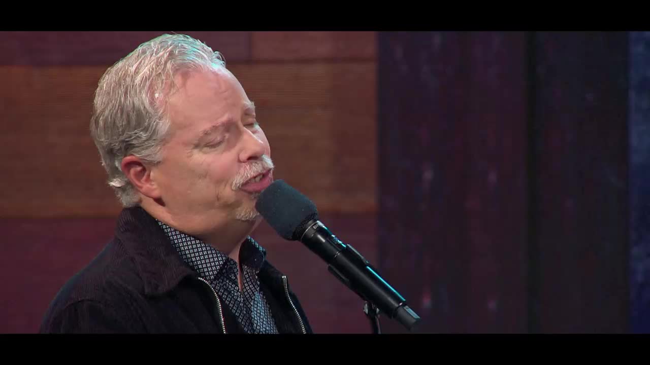 A man with graying hair and a mustache sings into a microphone, his eyes closed. He's performing on a stage, likely for a broadcast from the 3ABN Praise Him Music Network.