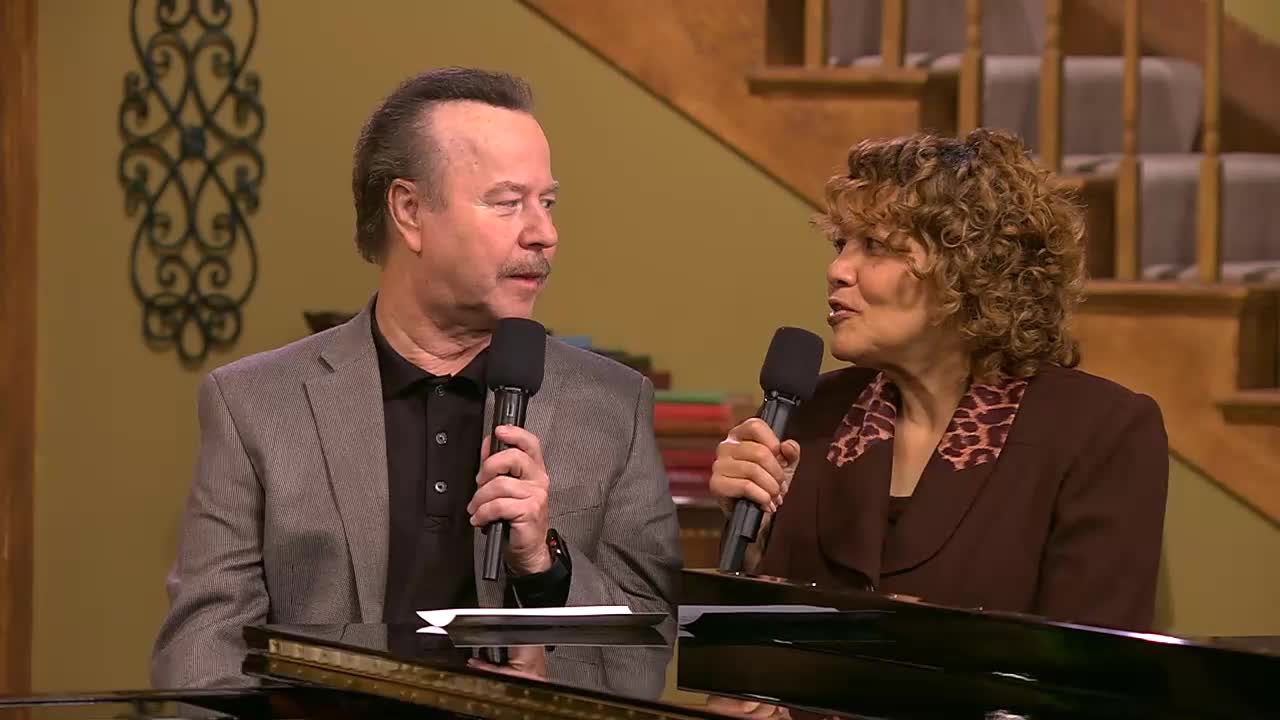 A man in a gray suit and a woman in a brown jacket are holding microphones. They appear to be in conversation, perhaps for a program on the 3ABN Praise Him Music Network.