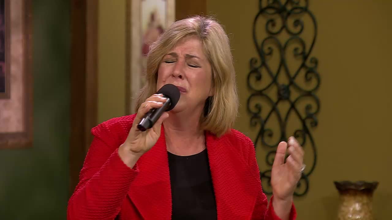 A woman in a bright red jacket sings with her eyes closed, holding a microphone to her lips. Her left hand is raised, fingers splayed, as if caught in a moment of passionate delivery.