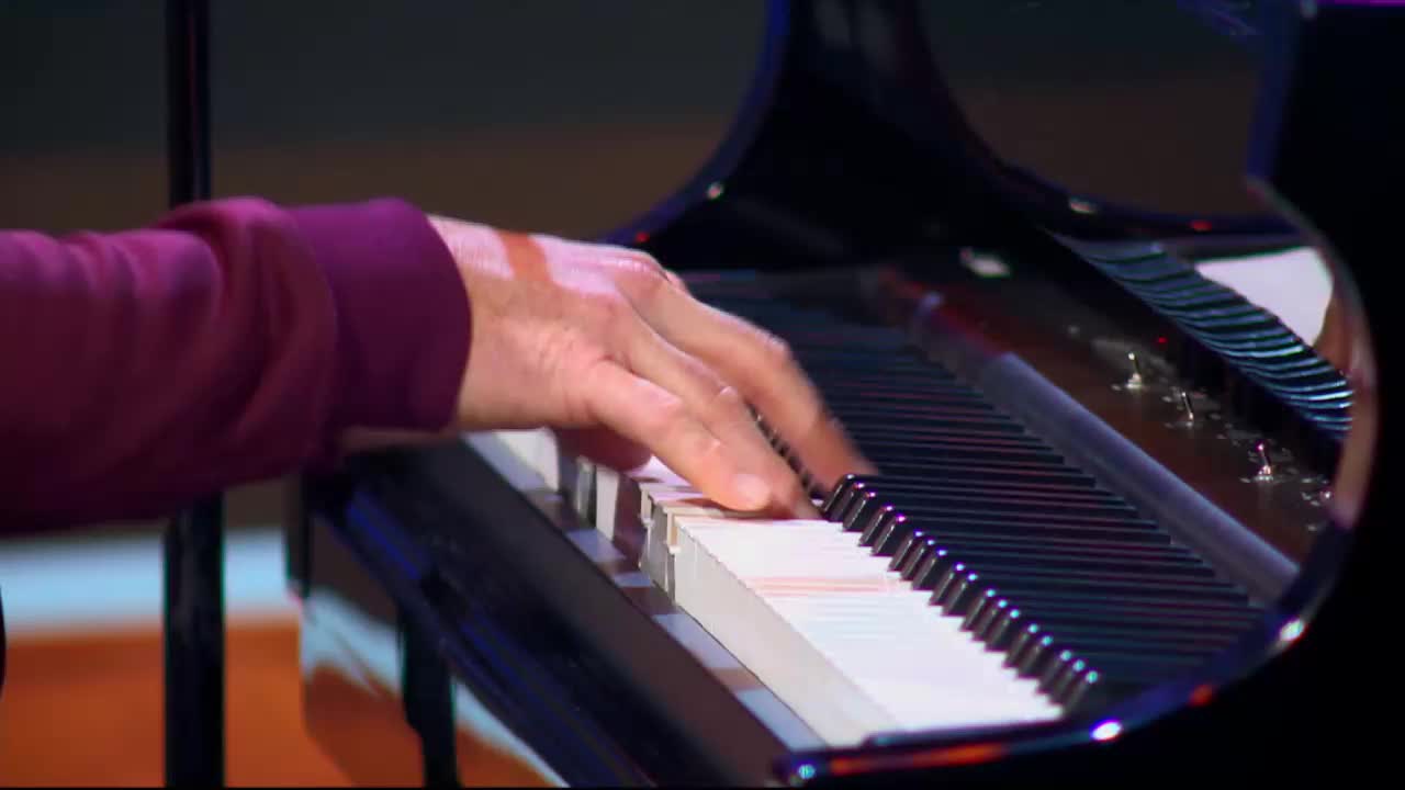 Fingers dance across the piano keys, their movements fluid and precise. The polished black surface of the grand piano reflects the stage lights, creating a dazzling display.