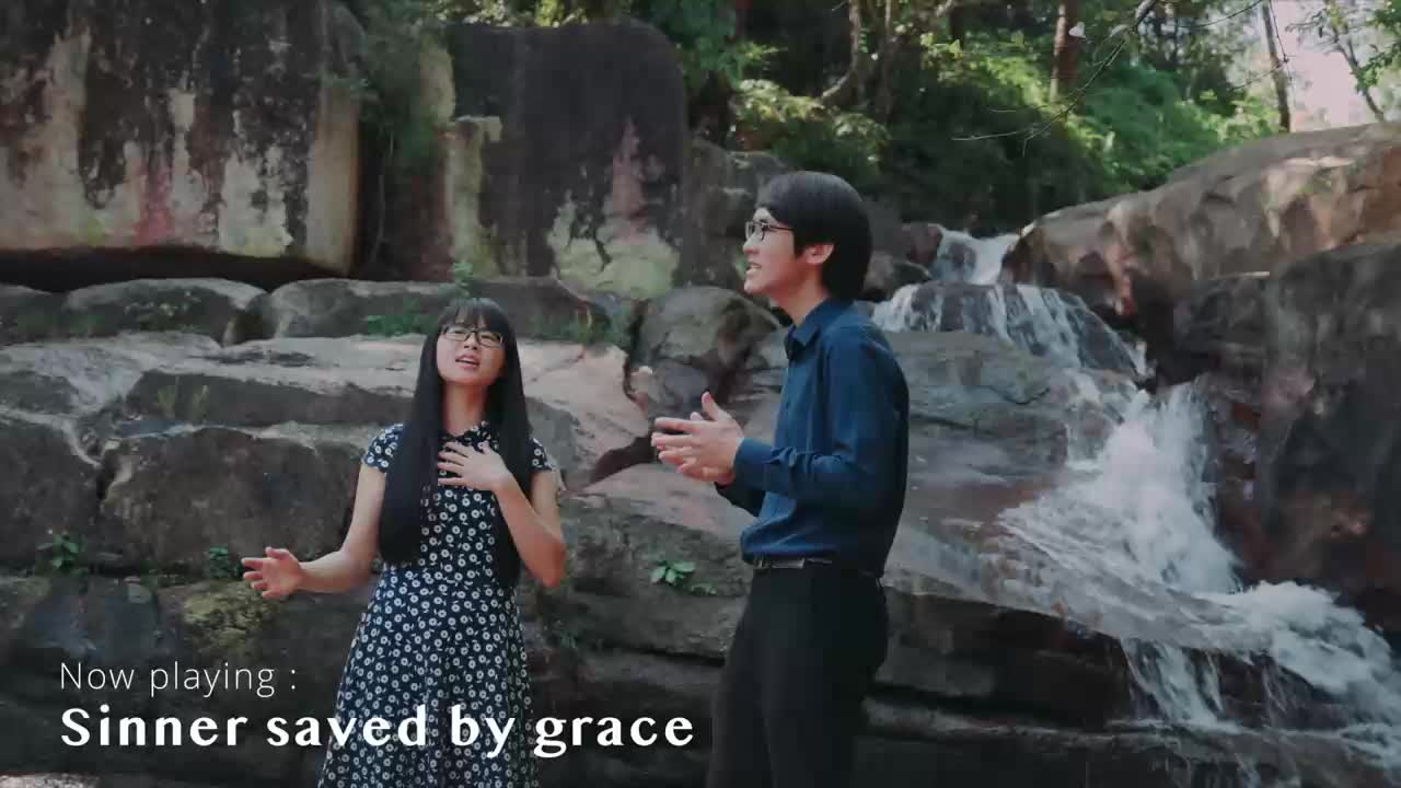 A young woman in a floral dress and a man in a blue shirt stand by a cascading waterfall. They are singing, their hands clasped or resting on their chests. The song playing is "Sinner saved by grace."