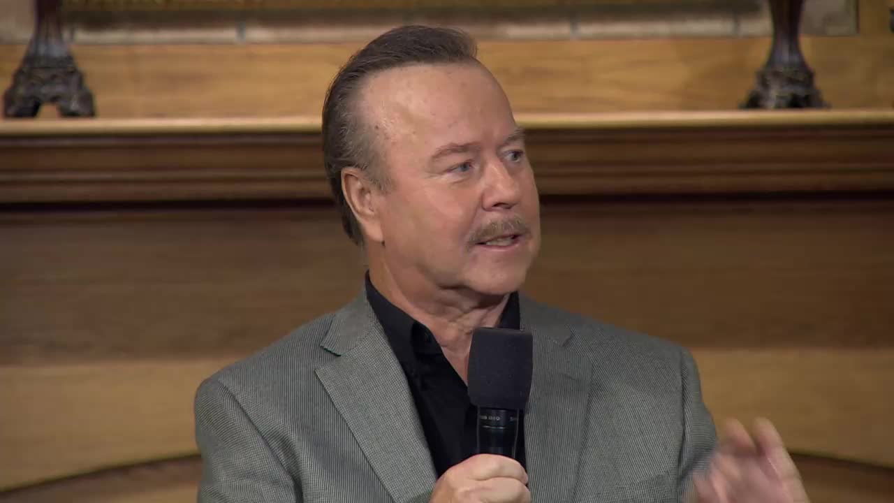 A man with a mustache speaks into a microphone, his right hand gesturing. He's dressed in a suit jacket over a black shirt, likely during a broadcast for the 3ABN Praise Him Music Network.