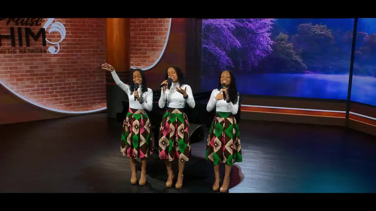 Three young women stand on a stage, singing into microphones. They wear matching white tops and vibrant, patterned skirts.