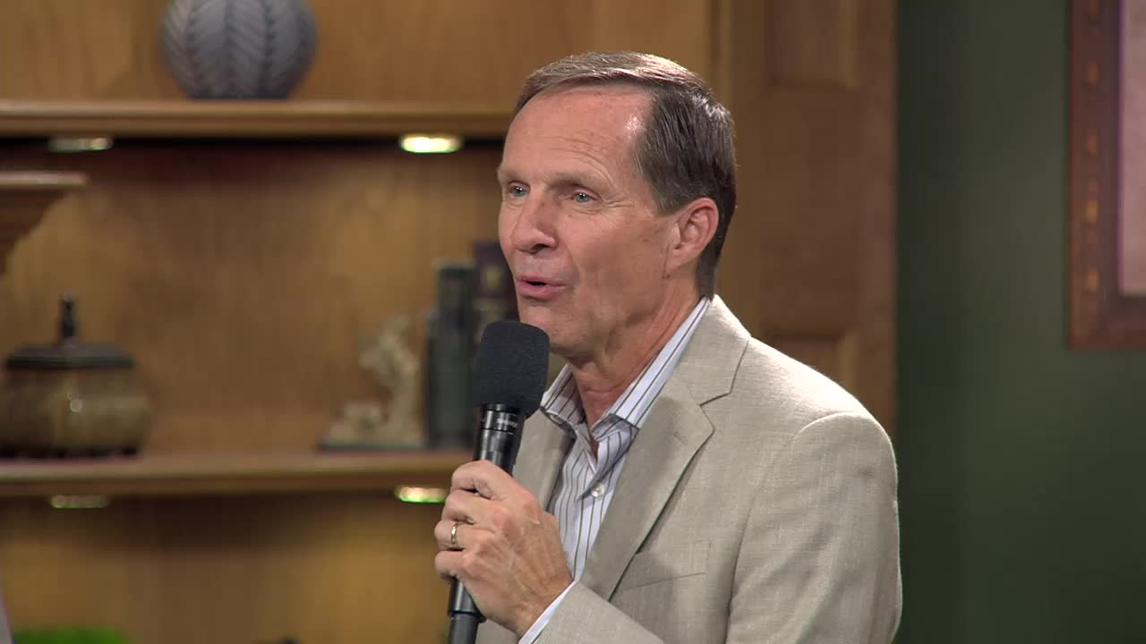 A man in a light-colored suit speaks into a microphone, his mouth open mid-sentence. He's on a set that looks like a home library, with shelves of books and decorative items behind him.