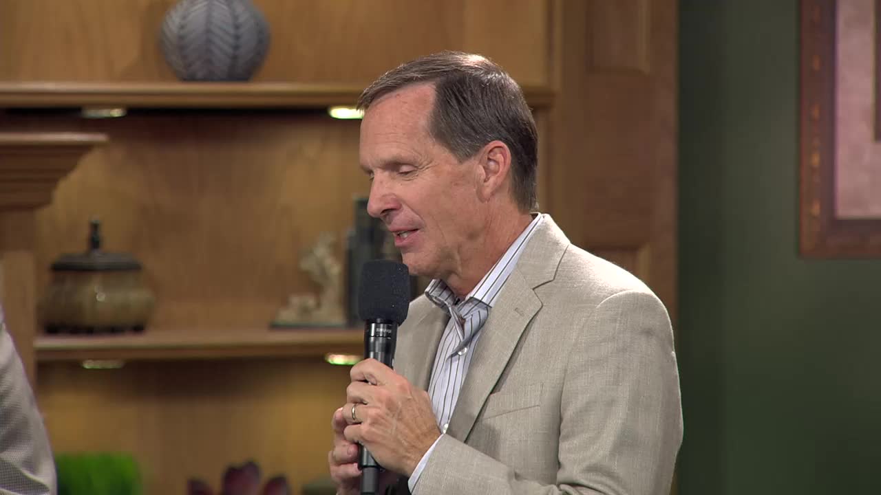 A man in a light-colored suit holds a microphone, his gaze directed downward. He's speaking into the mic, his expression thoughtful.