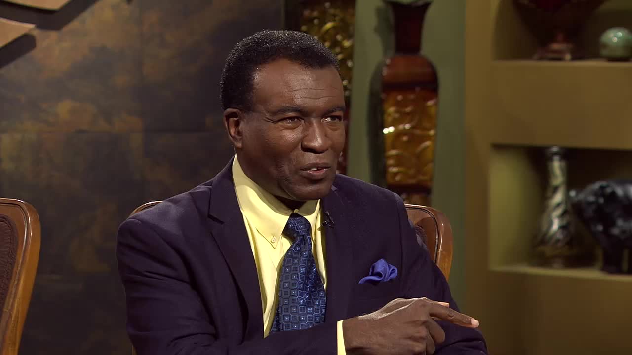 A man in a dark suit and yellow shirt gestures with his right hand while speaking. He's seated in a studio setting, likely for a program on the 3ABN Praise Him Music Network.