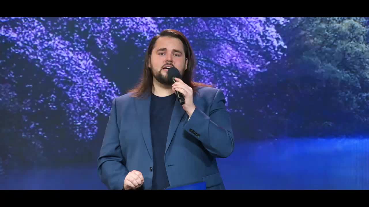 A man with long brown hair, wearing a blue suit jacket over a dark shirt, speaks into a microphone. Behind him, a projected backdrop displays a serene scene of trees and water in shades of purple and blue.