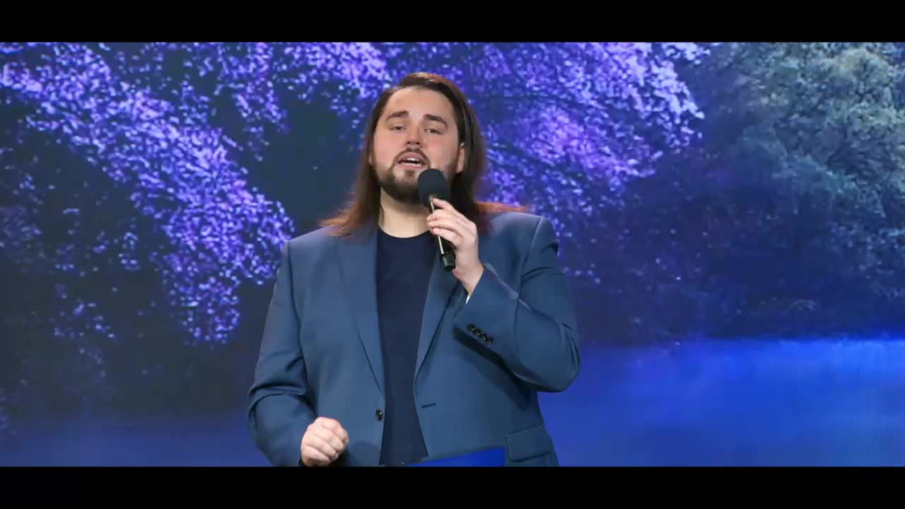 A man in a blue suit stands on a stage, holding a microphone. He's speaking into it, his left hand gesturing slightly. Behind him, a large screen displays a serene, blue-toned landscape with trees and water.
