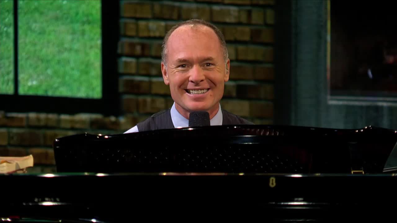 A man with a wide smile sits behind a grand piano, ready to play. He's dressed in a suit vest and tie, with a microphone in front of him.