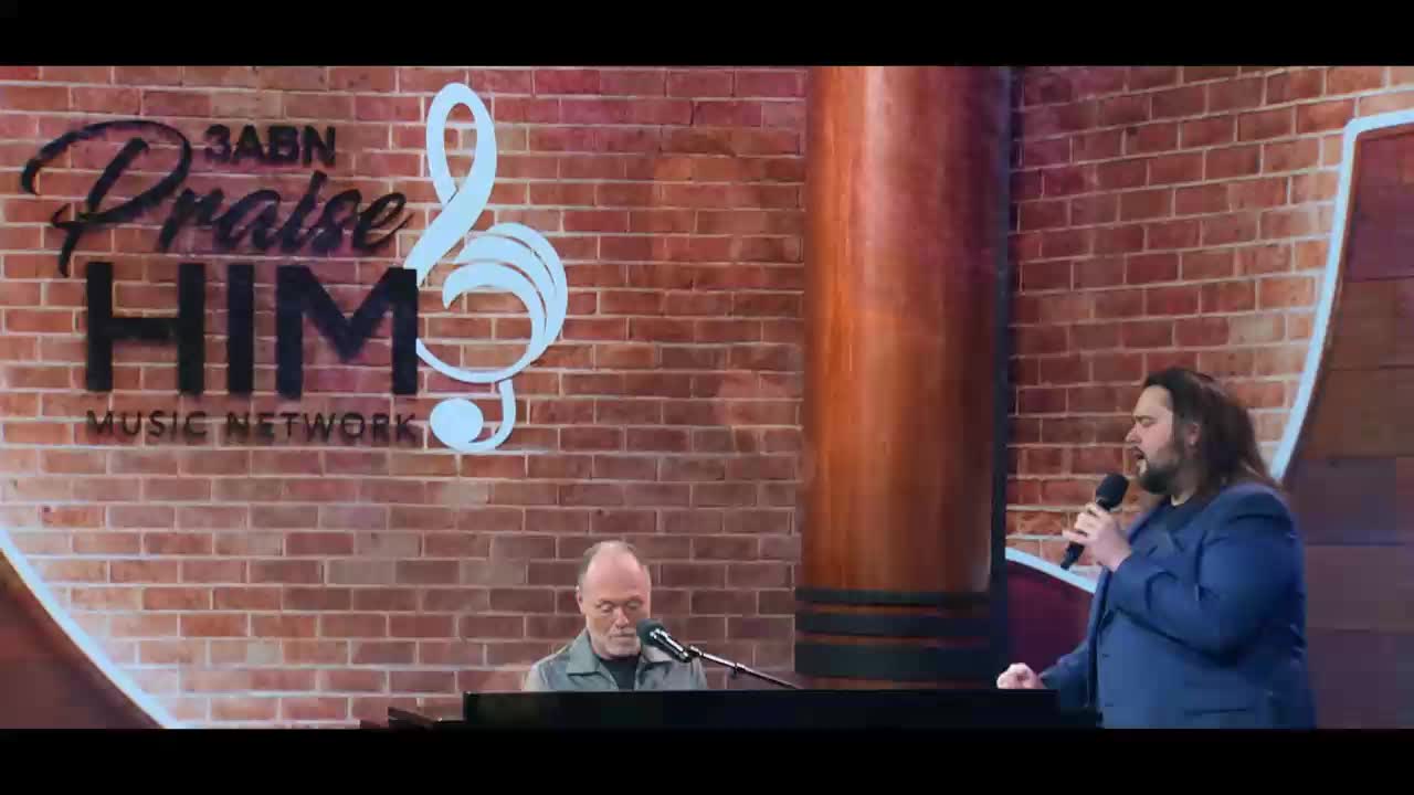 A man with long hair sings into a microphone while another man plays the piano. The 3ABN Praise Him Music Network logo hangs behind them on a brick wall.