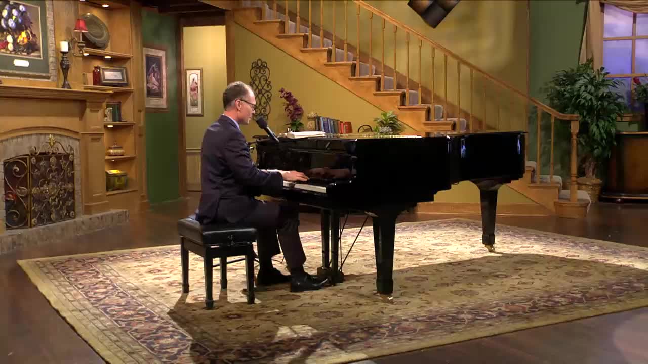 A man in a suit plays a grand piano on a rug in a studio setting. He's focused on the keys, his hands moving across them.