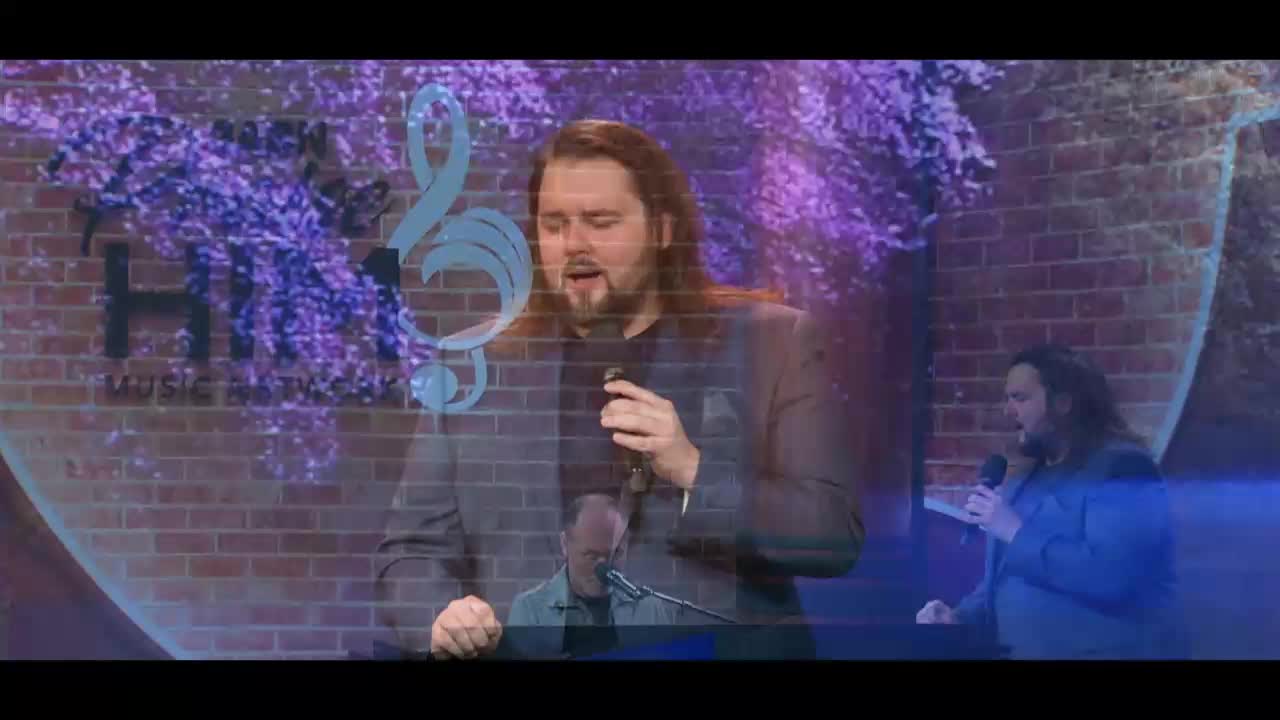 A man with long brown hair sings into a microphone, his eyes closed in concentration. Behind him, a brick wall is visible, overlaid with a large, stylized treble clef and a shimmering purple floral pattern.