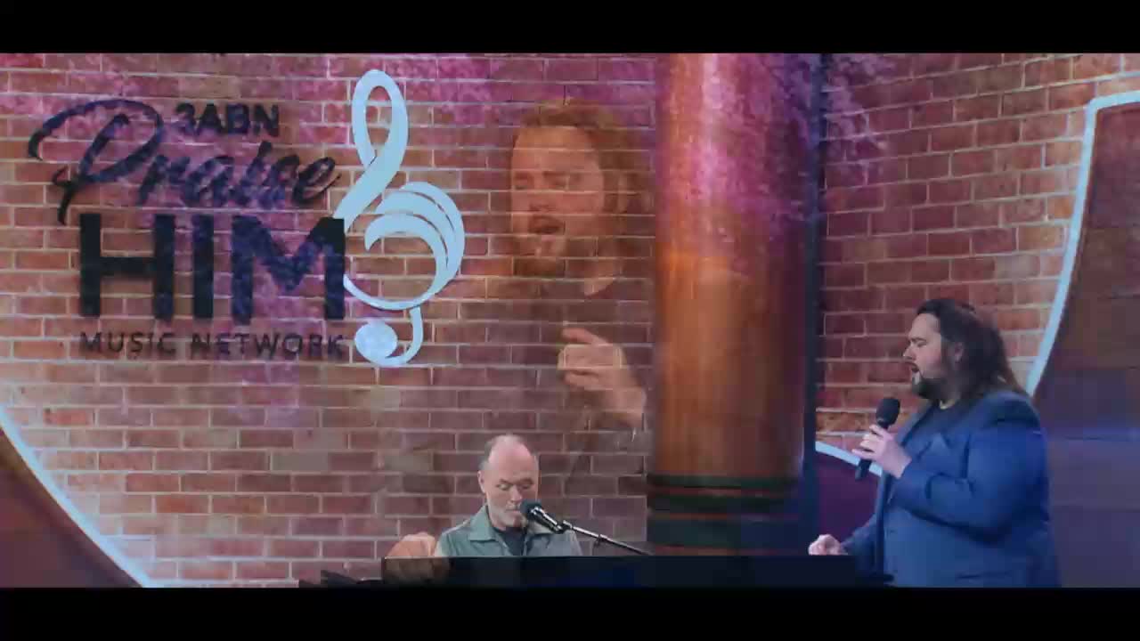 A man with long, dark hair stands holding a microphone and singing into it. To his left, another man plays the piano. Behind them, the 3ABN Praise Him Music Network logo is visible on a brick wall.