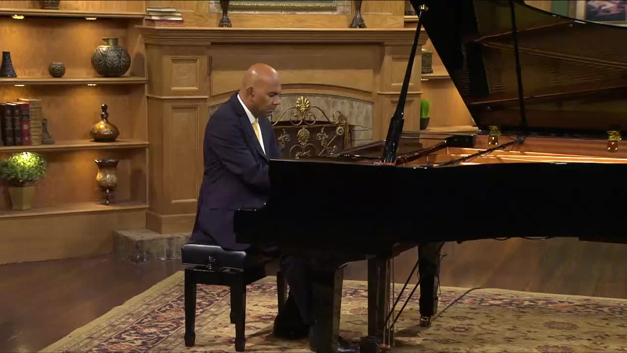 A man in a suit plays a grand piano, his hands moving across the keys. The scene is set in a warm, wood-paneled room, likely for a 3ABN Praise Him Music Network broadcast from the United States.