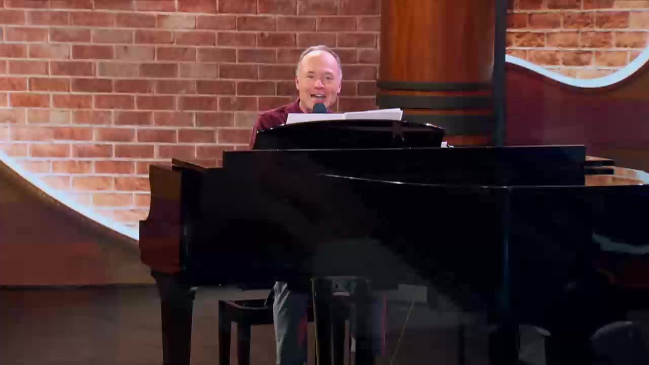 A man in a maroon shirt smiles as he plays a grand piano. He's on a stage with a brick backdrop, likely for a broadcast on the 3ABN Praise Him Music Network.