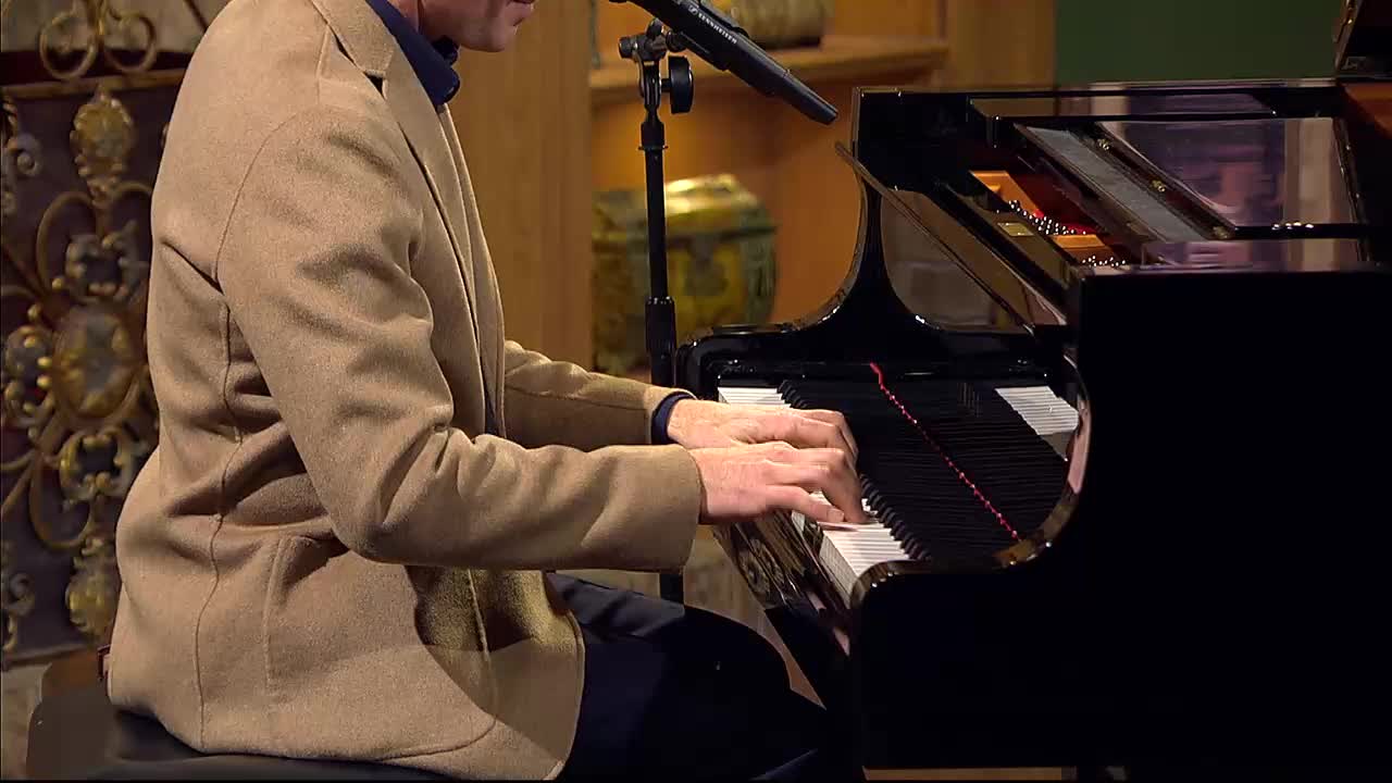 A man in a tan jacket plays a grand piano, his fingers moving across the keys. This performance is part of a 3ABN Praise Him Music Network broadcast from the United States.