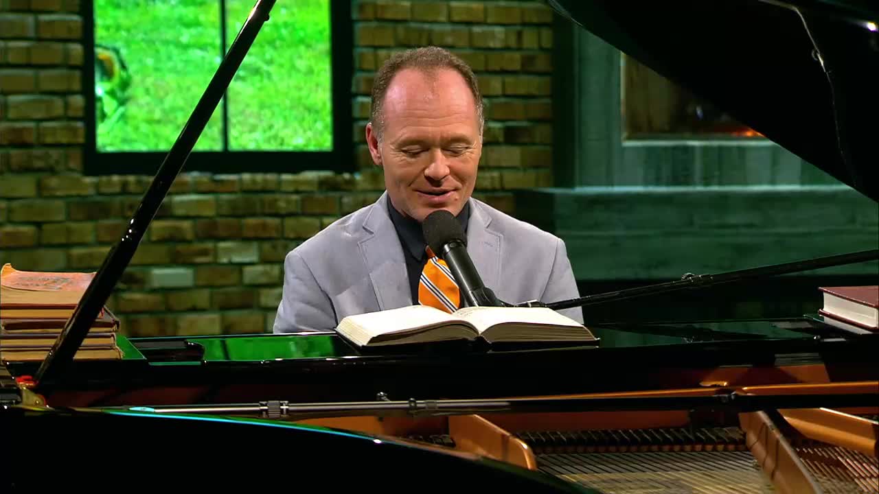 A man in a light suit and orange tie sits at a grand piano, his eyes closed as he reads from an open hymnal. He's about to play a song for a 3ABN Praise Him Music Network broadcast.