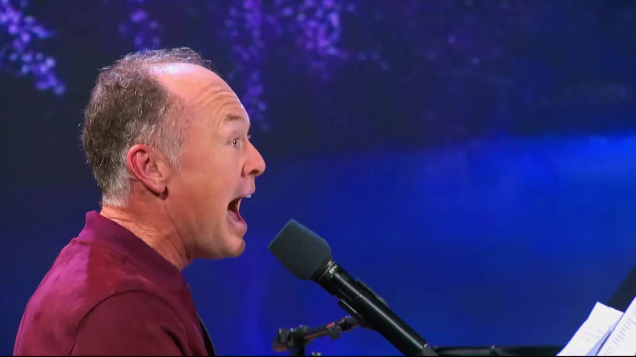 A man with a maroon shirt belts out a song into a microphone, his mouth wide open. He's performing on a stage with a dark blue backdrop, likely for a 3ABN Praise Him Music Network broadcast.