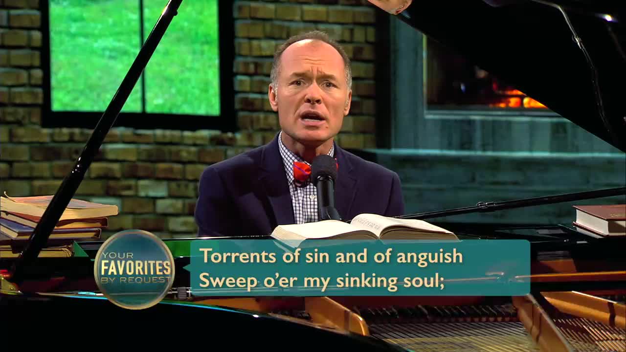 A man in a suit and bowtie stands before a microphone, singing from an open hymnal. The lyrics, "Torrents of sin and of anguish Sweep o'er my sinking soul," appear on a screen.