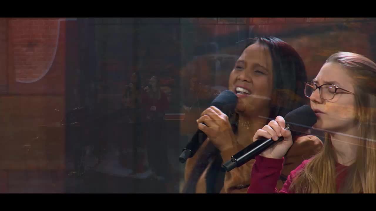 Two women sing into microphones, their voices blending in harmony. Behind them, a blurred background hints at a live performance for the 3ABN Praise Him Music Network.