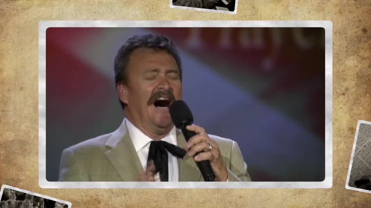 A man with a mustache belts into a microphone, his eyes closed in passionate song. He's dressed in a light suit jacket and a black bolo tie, performing on a stage lit with red and blue.