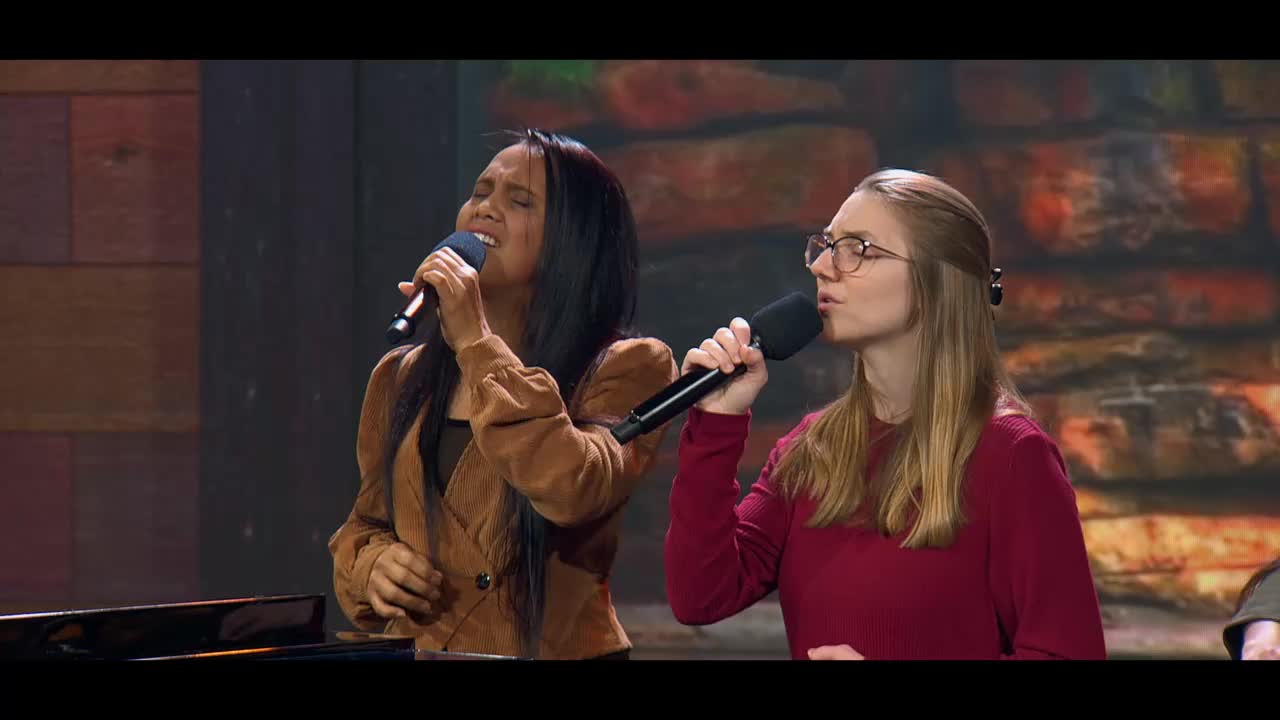 Two women stand at microphones, singing with their eyes closed. The woman on the left wears a brown jacket, while the one on the right has on a maroon sweater.