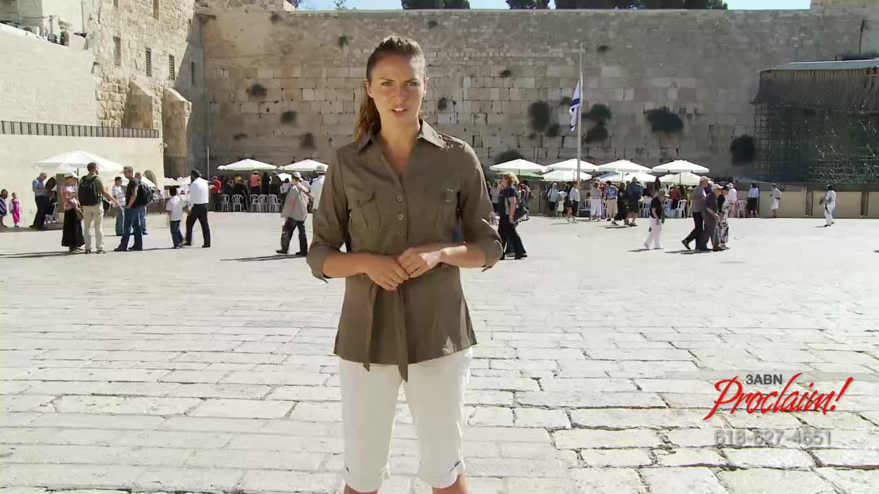 A woman in a khaki shirt and white capri pants stands in front of a large stone wall, speaking to the camera. People stroll across the sun-drenched plaza behind her, with the 3ABN Proclaim! logo visible in the corner.
