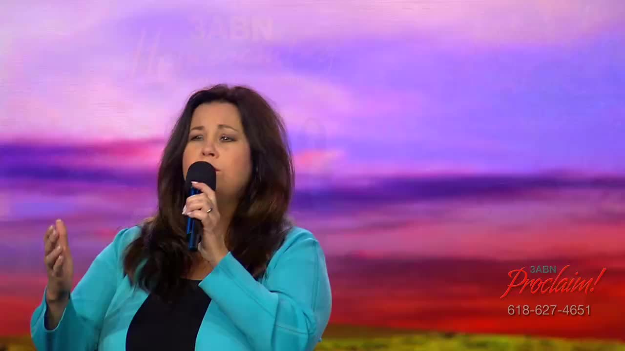 A woman in a teal jacket sings into a microphone, gesturing with her left hand. The background is a gradient of purple, pink, and orange, with the 3ABN Proclaim! logo visible.
