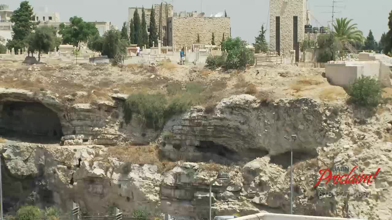 A rocky hillside, dotted with small trees and shrubs, slopes down towards a cave opening. In the distance, a stone building and a cemetery are visible under a bright sky, as seen on 3ABN Proclaim! Network.

