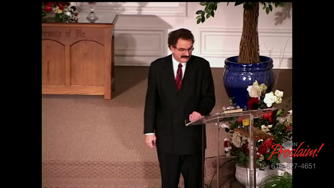 A man in a dark suit stands at a clear podium, speaking. The 3ABN Proclaim! Network logo is visible in the corner, and a floral arrangement sits nearby.
