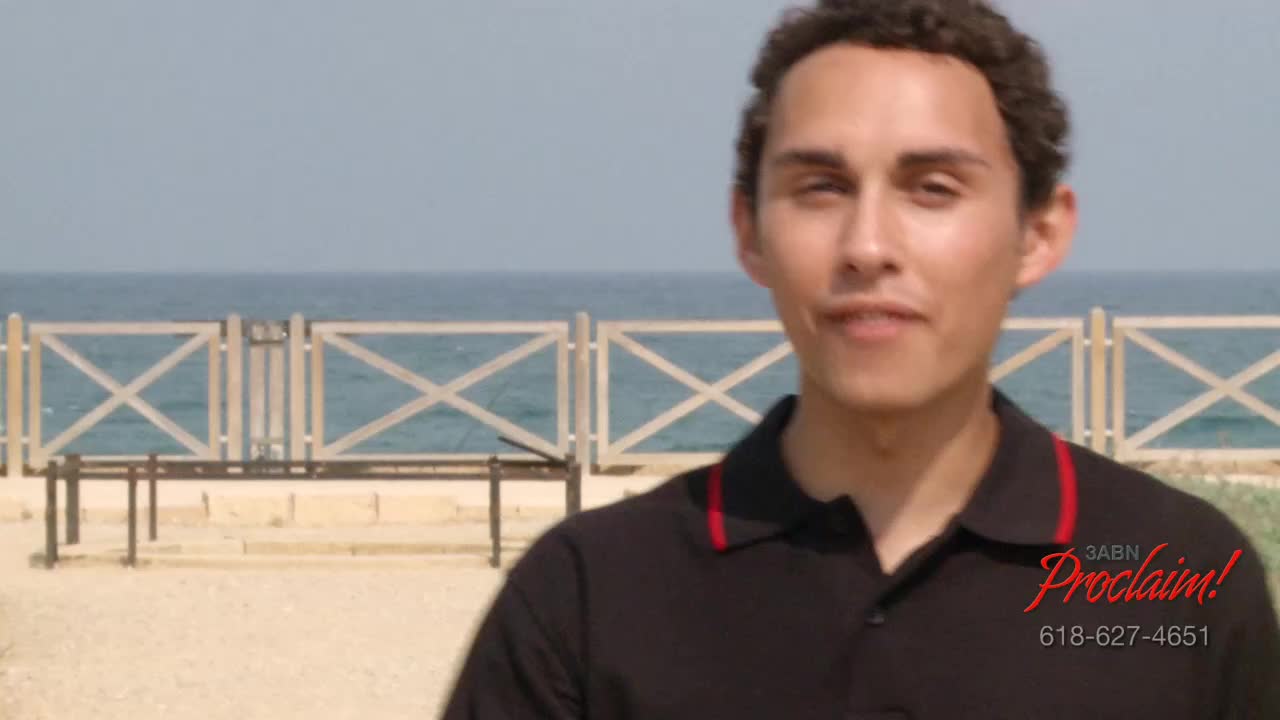 A man in a black shirt with red trim smiles at the camera, the 3ABN Proclaim! logo visible in the corner. Behind him, a wooden fence and the ocean are visible under a bright sky.
