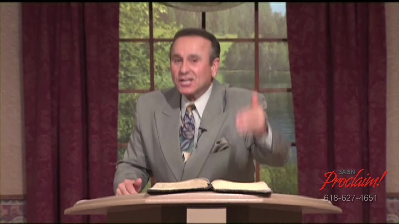 A man in a gray suit gestures emphatically while speaking, his words likely broadcast across the United States on the 3ABN Proclaim! Network. An open book rests on the lectern before him.
