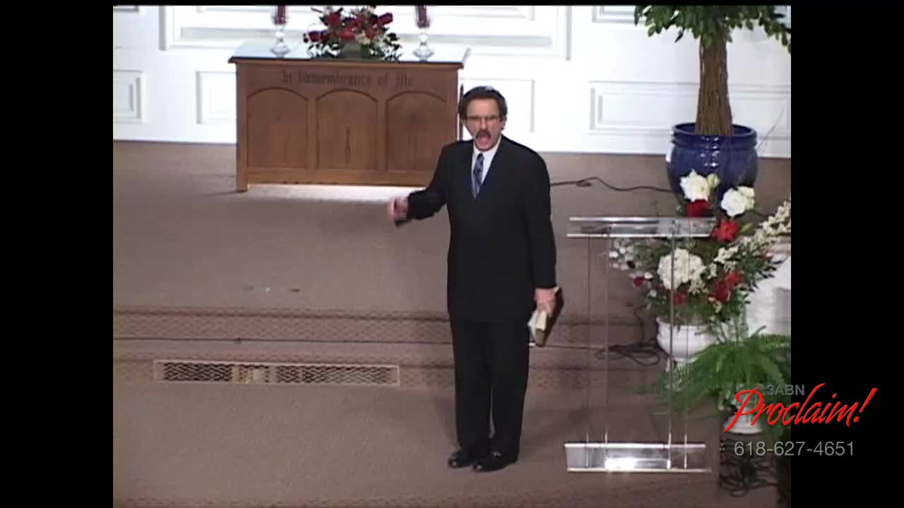 A man in a dark suit gestures with his right hand while holding a book in his left. He stands on the stage of a church, likely delivering a sermon for the 3ABN Proclaim! Network.
