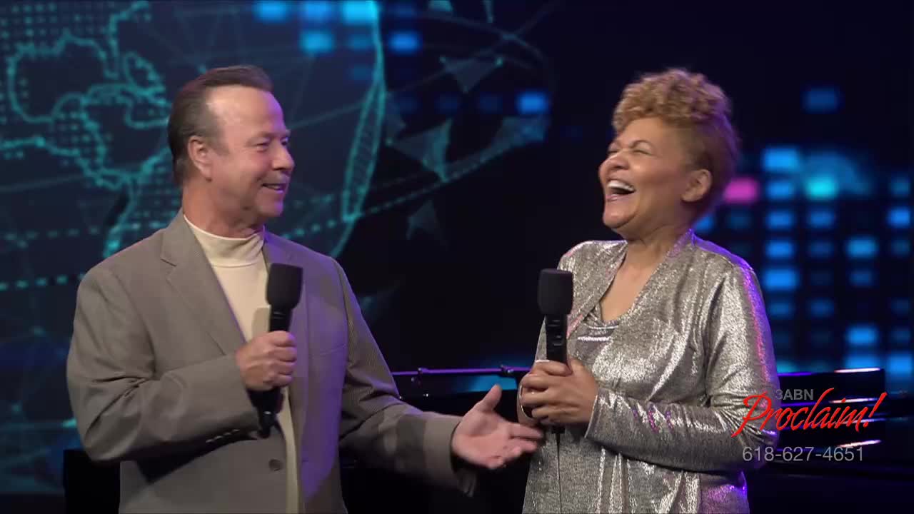 A man in a gray suit gestures towards a woman in a shiny silver jacket, both holding microphones. The woman is laughing heartily, her head thrown back.
