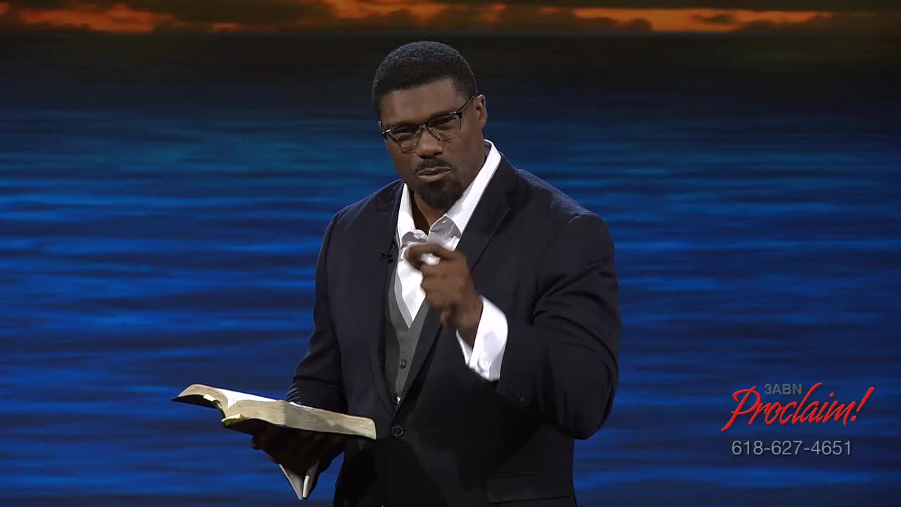 The man in the suit, holding a book, is pointing directly at the camera. He's speaking on the 3ABN Proclaim! Network, in the United States.
