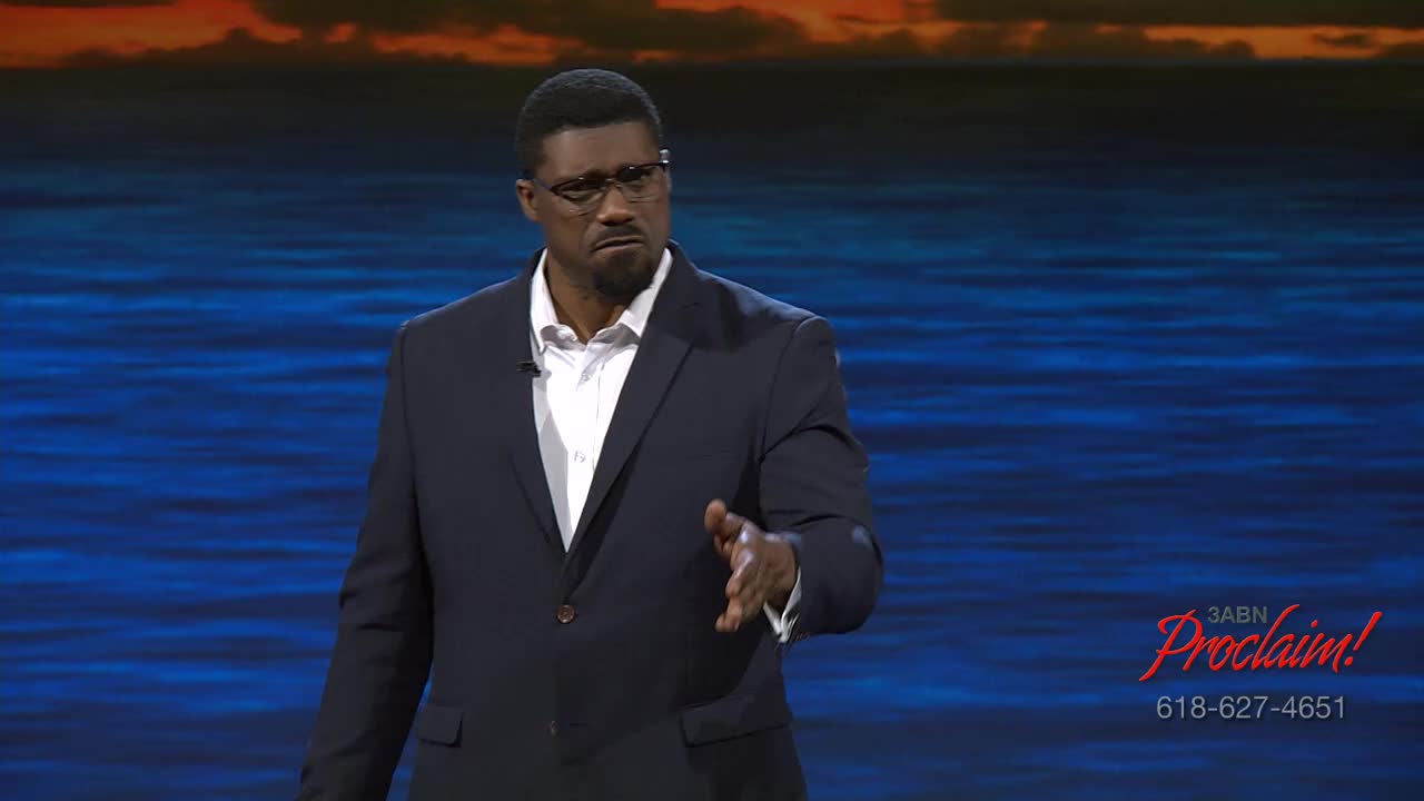 A man in a navy suit gestures with his right hand, speaking on a stage in front of a backdrop of water and sky. The 3ABN Proclaim! logo is in the bottom right corner.
