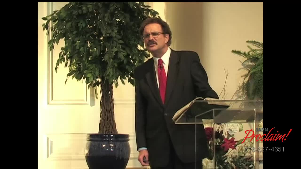 A man in a suit, with a red tie, stands at a clear podium, speaking. Behind him, a large potted tree and the logo for 3ABN Proclaim! are visible.
