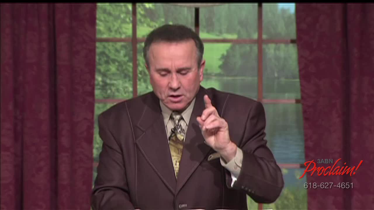 The man in the brown suit gestures with his right index finger as he speaks. Behind him, a window offers a view of trees and a lake, while the 3ABN Proclaim! logo is visible in the corner.
