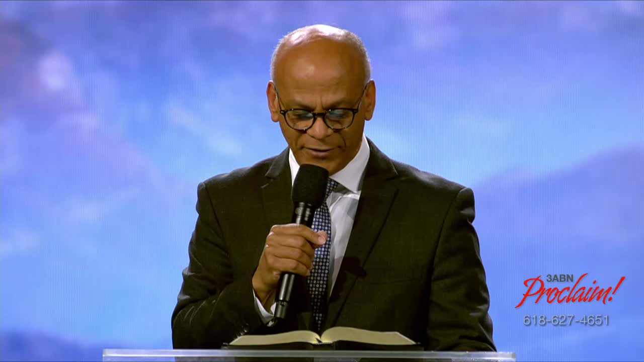 A man in a dark suit, glasses, and tie stands behind a lectern, reading from an open book. He holds a microphone in his right hand, and the 3ABN Proclaim! logo is visible on the right.
