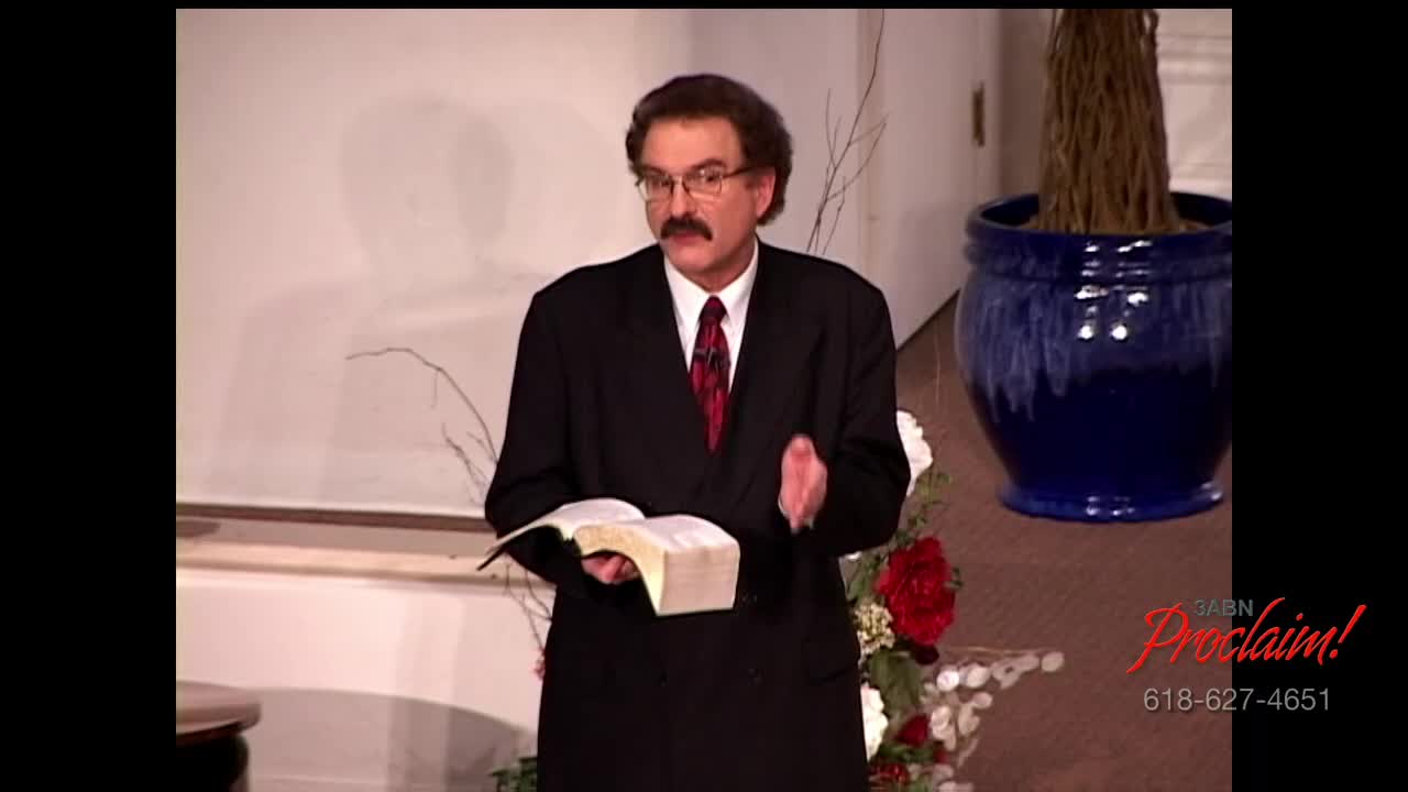 A man in a suit stands holding a book, gesturing with his hands. He's speaking on a set for 3ABN Proclaim!, a television ministry in the United States.
