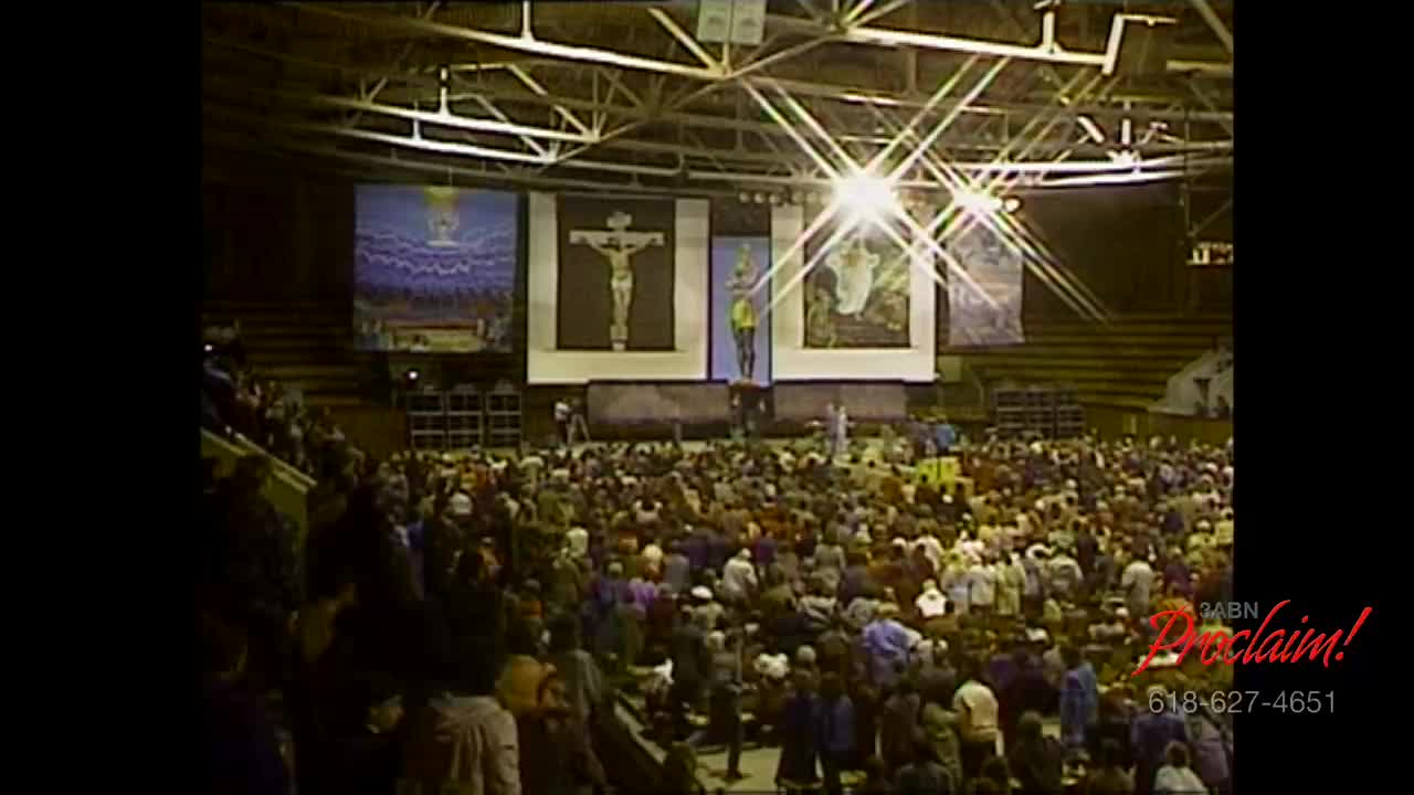 A large crowd fills the arena, facing a stage with religious imagery and bright lights. People are standing and moving about, likely waiting for the next segment of the 3ABN Proclaim! Network broadcast.
