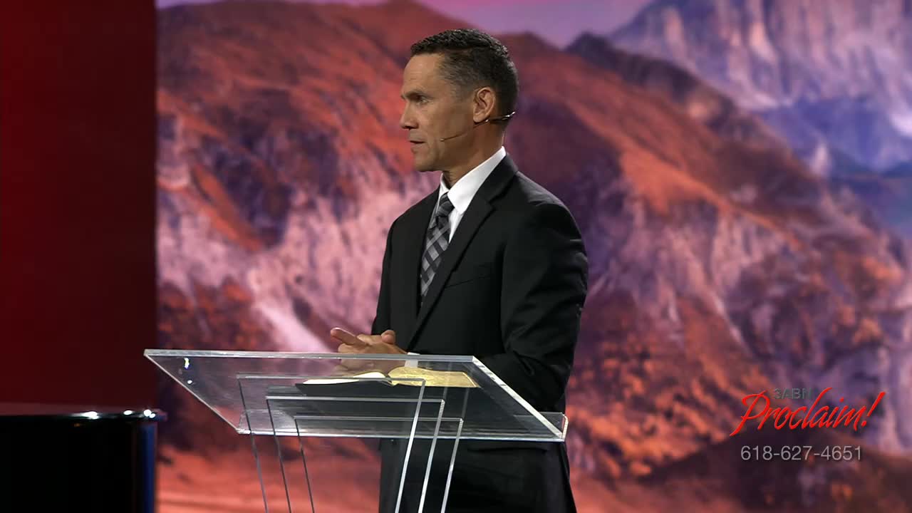 A man in a suit stands behind a clear podium, speaking with his hands clasped. Behind him, a backdrop of a mountain range is visible on the 3ABN Proclaim! Network.
