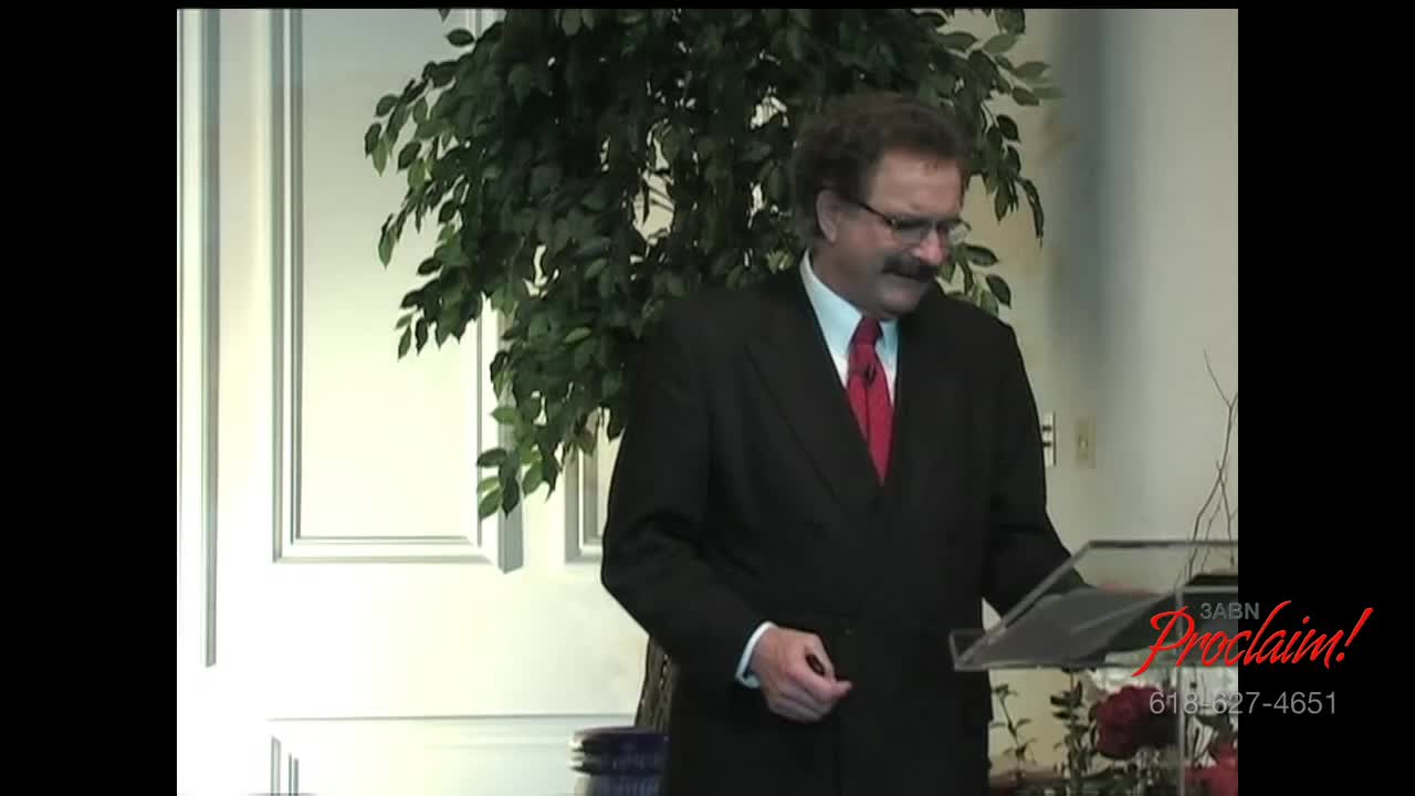 A man in a suit and red tie stands behind a podium, looking down at his notes. Behind him, a large potted plant and the 3ABN Proclaim! Network logo are visible.
