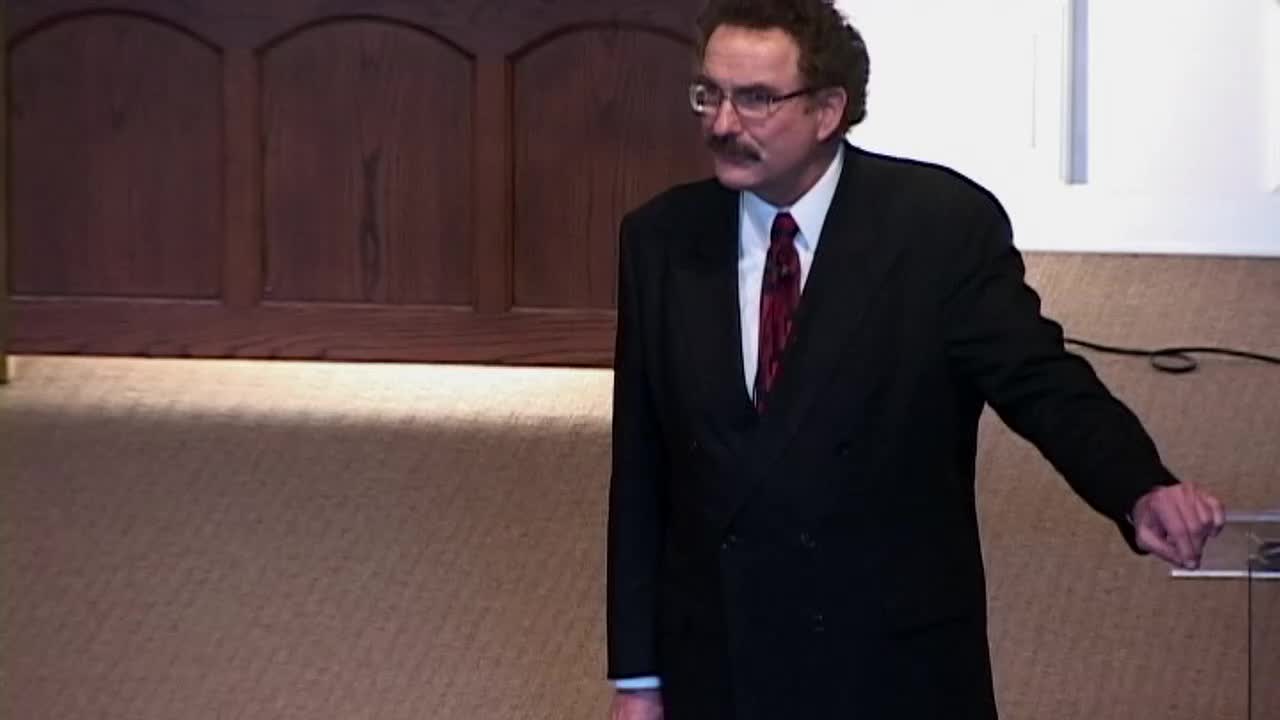 A man in a dark suit and glasses stands near a lectern, gesturing with his right hand. He's speaking on the 3ABN Proclaim! Network, likely in the United States, with a wooden backdrop behind him.
