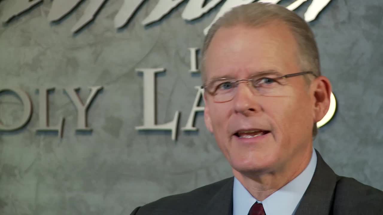 A man in a suit and glasses speaks, his mouth open as he addresses the camera. Behind him, the words "Holy Land" are visible on a textured wall.
