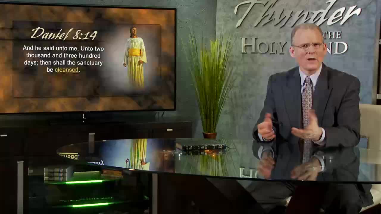 A man in a suit gestures with both hands, speaking at a desk in a television studio. Behind him, a screen displays a Bible verse and an image of Jesus, likely for a program on the 3ABN Proclaim! Network in the United States.
